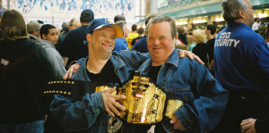 Two Noble participants smile and laugh while holding a large championship belt.