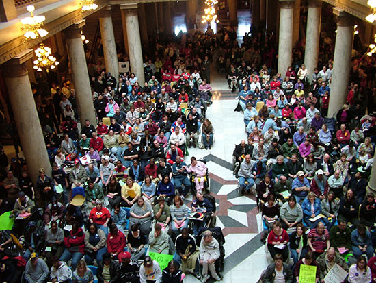 Large crowd of individuals as seen from above in a large columned room.